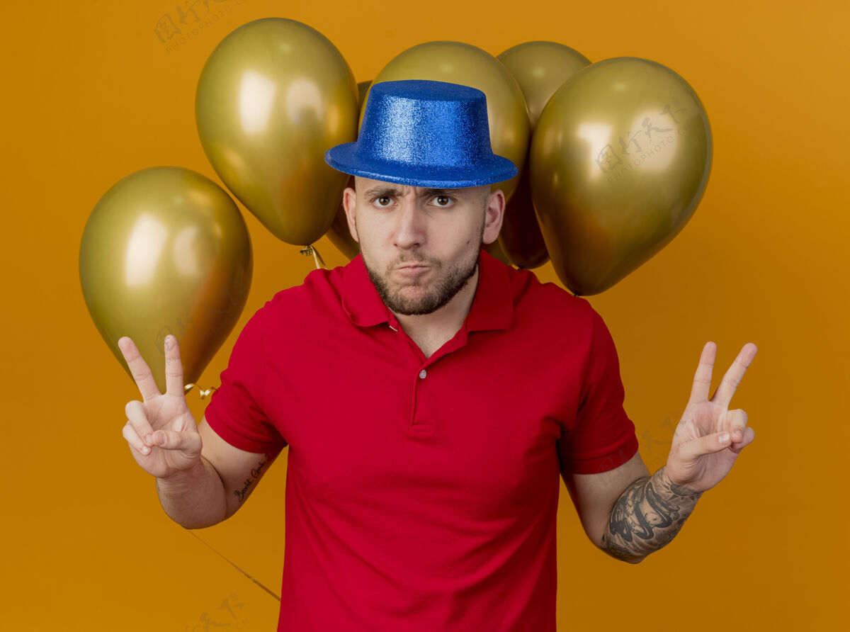 气球皱眉的年轻帅哥戴着派对帽站在气球前看着前面做着和平手势孤立在橙色的墙上英俊立场帽子
