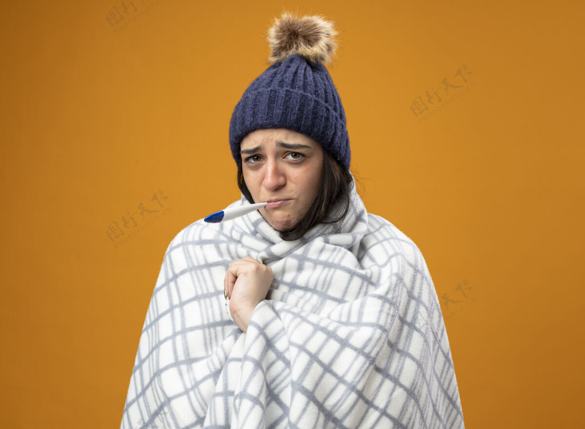 人穿着长袍 戴着格子布冬帽的虚弱的年轻病妇抓着格子布看着前面 嘴里叼着温度计 隔离在橙色的墙上长袍疾病冬天