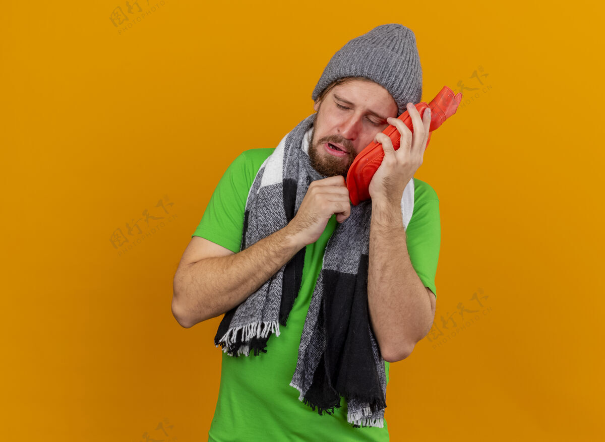 衣服一个戴着冬日帽子 戴着围巾 拿着热水袋 闭着眼睛 在橘色的墙上摸着脸的年轻帅哥站着热帽子