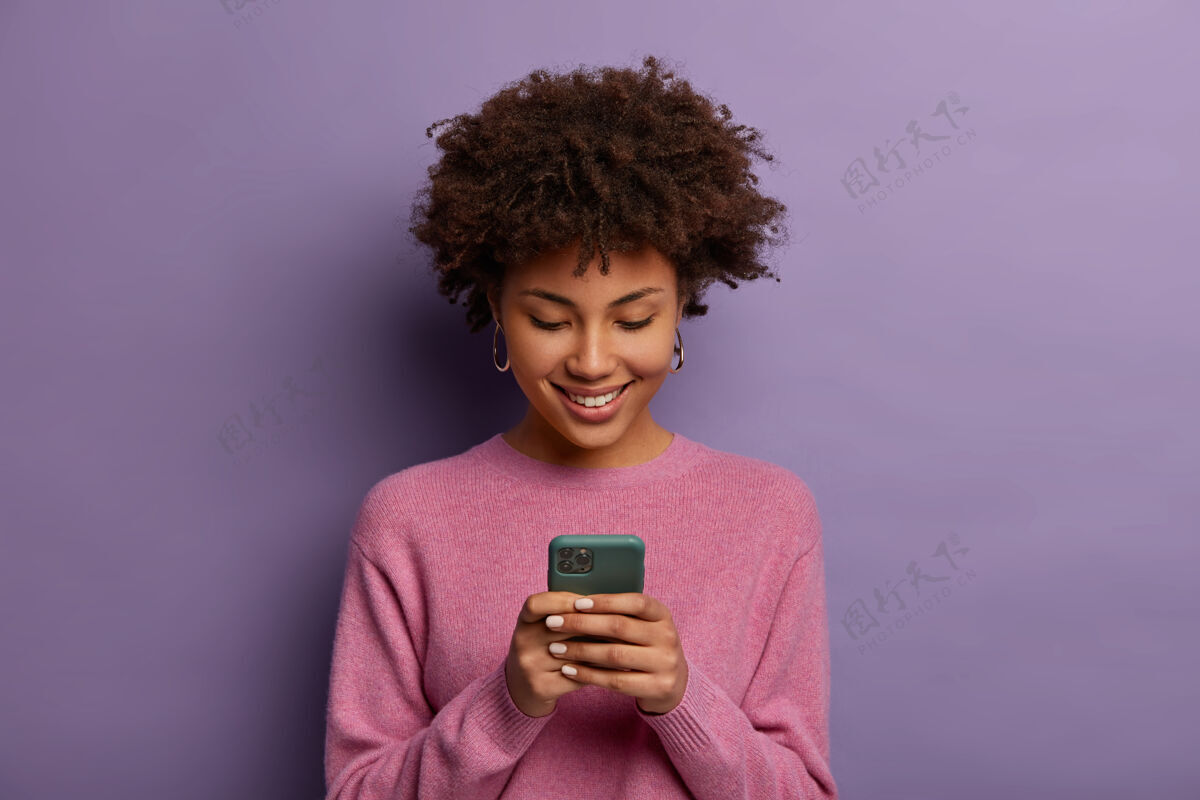 聊天漂亮的卷发女人手持现代手机 在智能手机上输入信息 享受在线交流 下载聊天专用应用程序 温柔微笑 孤立在紫色的墙上满意短信脸