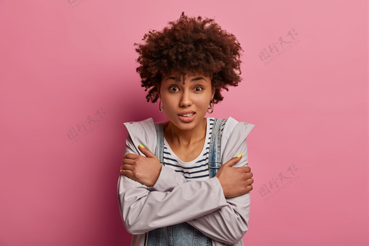 黑发卷曲的年轻非裔美国妇女的照片双臂交叉在身上 需要热身 在刮风的天气里冻僵 站着不安全和害怕 感觉寒冷和一文不值 对着粉色的墙壁摆姿势害怕室内消极