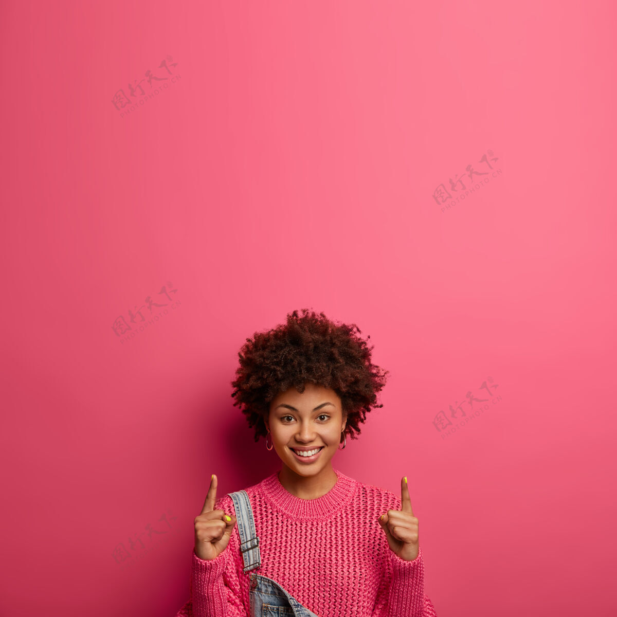 编织你的标志在这里高兴的微笑着的女人与非洲裔头发点和推广的东西向上 说遵循这个方向 穿针织毛衣 隔离在粉红色的墙壁营销和广告建议休闲促销