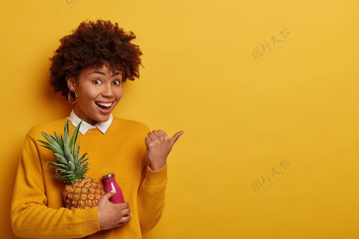 女性令人惊奇的高兴的女人有愉快的心情 拿着新鲜的菠萝和一瓶冰沙 把拇指放在一边 坚持节食 吃富含维生素的水果 穿黄色的衣服水果新鲜咯咯笑