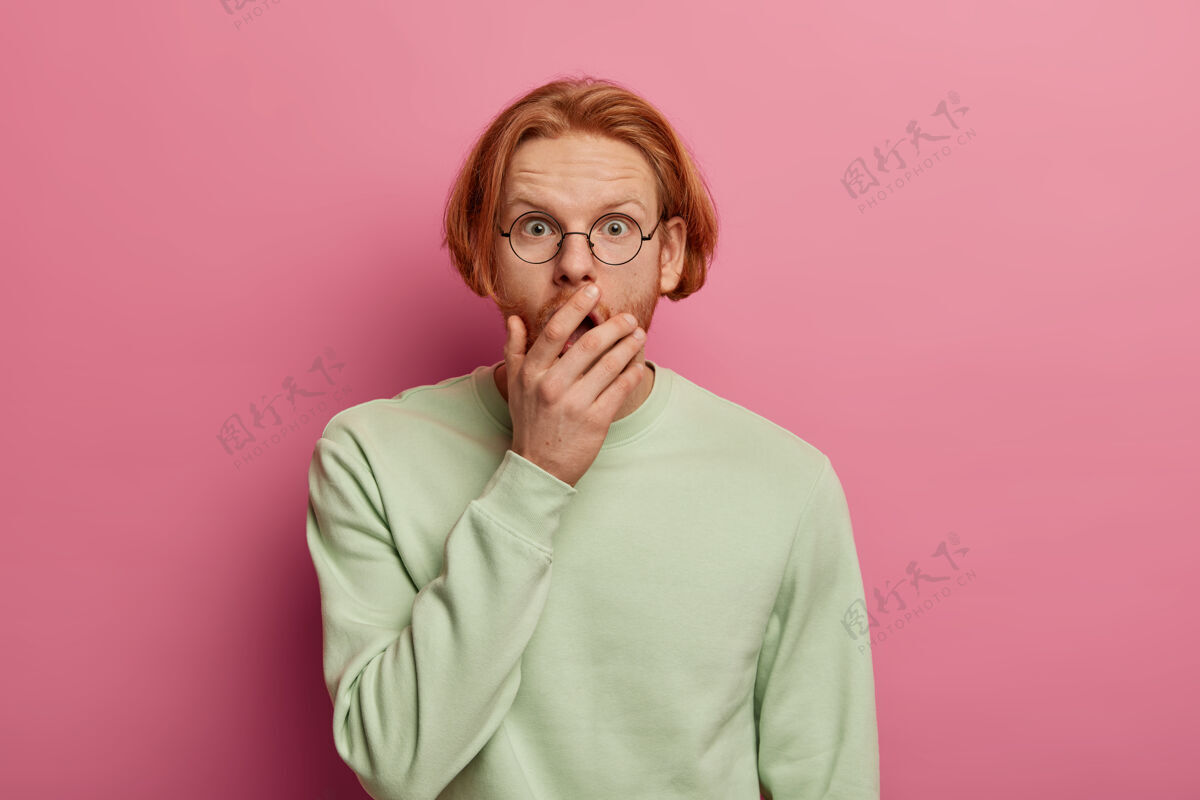 穿着一个孤立的镜头 惊愕的胡须 红发的时髦男人捂着张嘴 不敢相信令人震惊的消息 看到一些难以置信的事情 穿着休闲套头衫 对着粉色的墙壁摆姿势眼镜昏迷无语