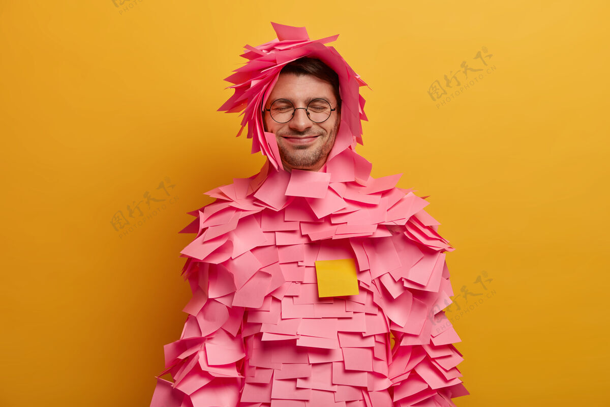 笔记积极的家伙在身体和头部贴上粉红色的胶纸 用胶纸做创意服装 戴眼镜 在办公室工作 隔离在黄色的墙上 闭上眼睛胡茬姿势眼镜