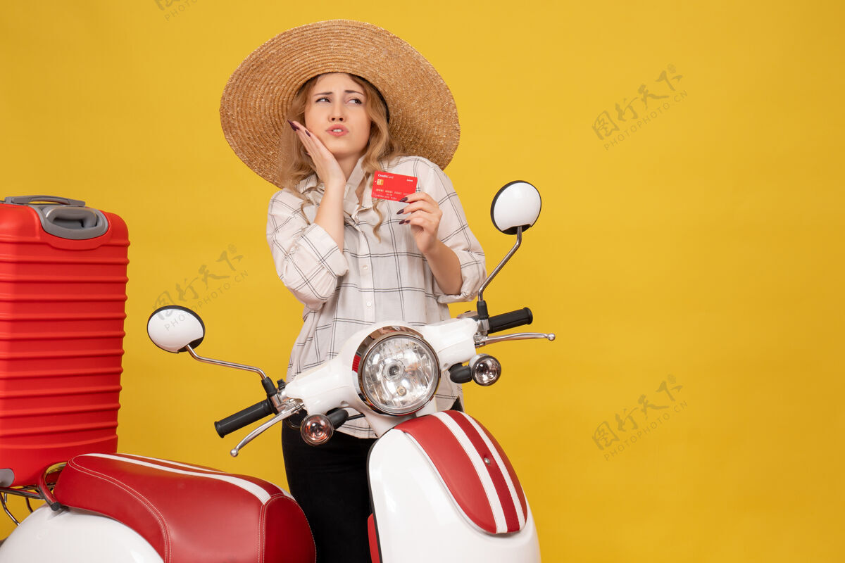 年轻女子俯视图：好奇的年轻女子戴着帽子坐在摩托车上拿着银行卡拿着行李人摩托车银行