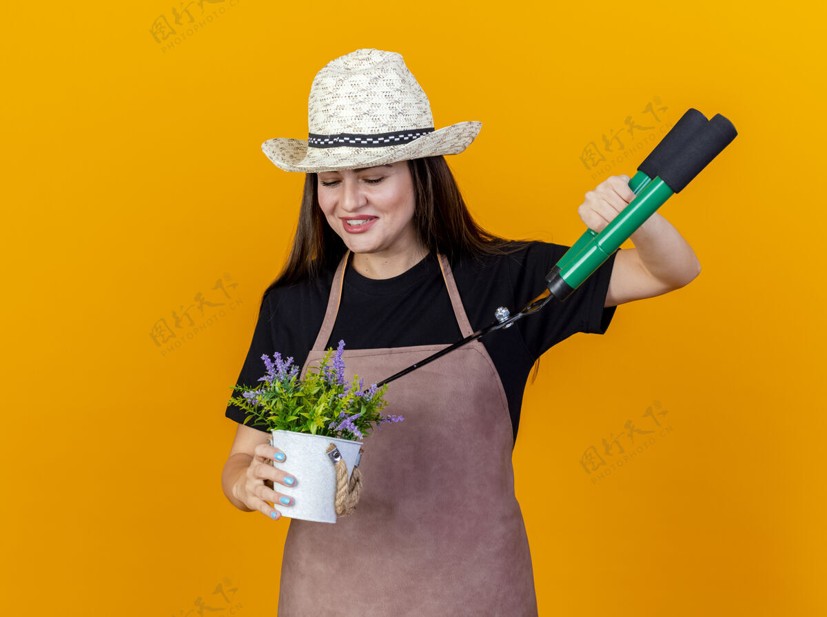 女孩微笑美丽的园丁女孩穿着制服 戴着园艺帽 拿着剪子在花盆里切花 剪子孤立在橙色的背景上微笑花盆花