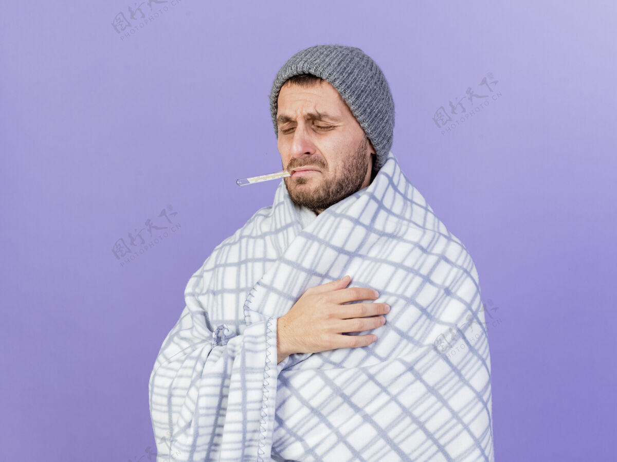 格子布闭上眼睛后悔的年轻人戴着冬天的帽子 围巾裹在格子布里 嘴里叼着温度计 隔离在紫色的背景上持有嘴巴围巾