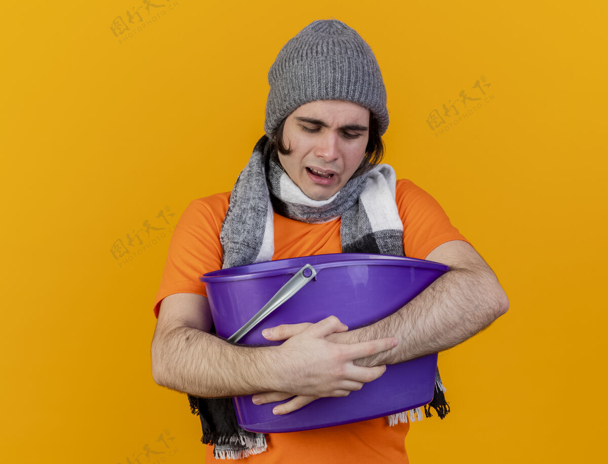 年轻体弱多病的年轻人戴着冬天的帽子 戴着围巾 手上拿着塑料桶 在橙色的背景上孤立着 恶心帽子水桶围巾