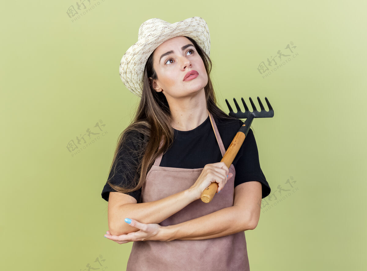 园艺抬头一看 高兴的美丽的园丁女孩穿着制服 戴着园艺帽 拿着耙子孤立在橄榄绿的背景上园艺耙子制服