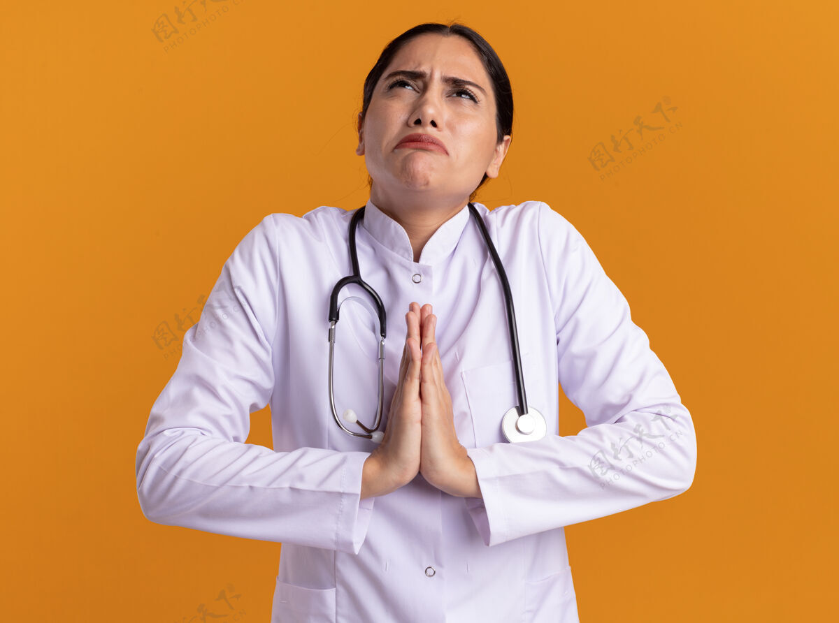 女人身穿医用外套的年轻女医生 脖子上戴着听诊器 双手合十 像在祈祷一样 站在橙色的墙上 带着希望的表情仰望着天空站着听诊器医生