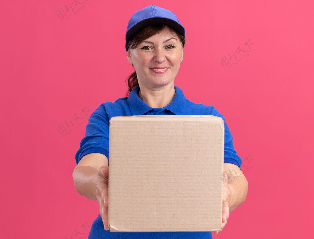 盒子身穿蓝色制服 头戴鸭舌帽的中年送货妇女站在粉红色的墙上 面带微笑 自信地看着前面的纸板箱送货女人年龄