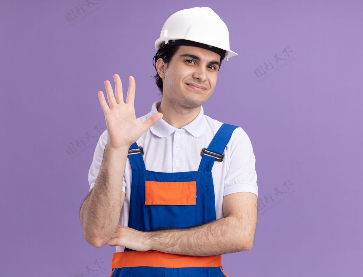 安全年轻的建筑工人穿着建筑制服 戴着安全帽 面带微笑 站在紫色的墙上挥手致意站着手建筑工人