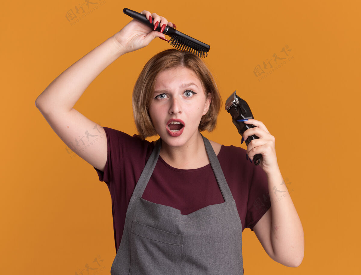 年轻年轻漂亮的女理发师站在橘色的墙上 围着围裙拿着剪子和梳子梳着头发 显得迷茫和惊讶理发师拿着漂亮
