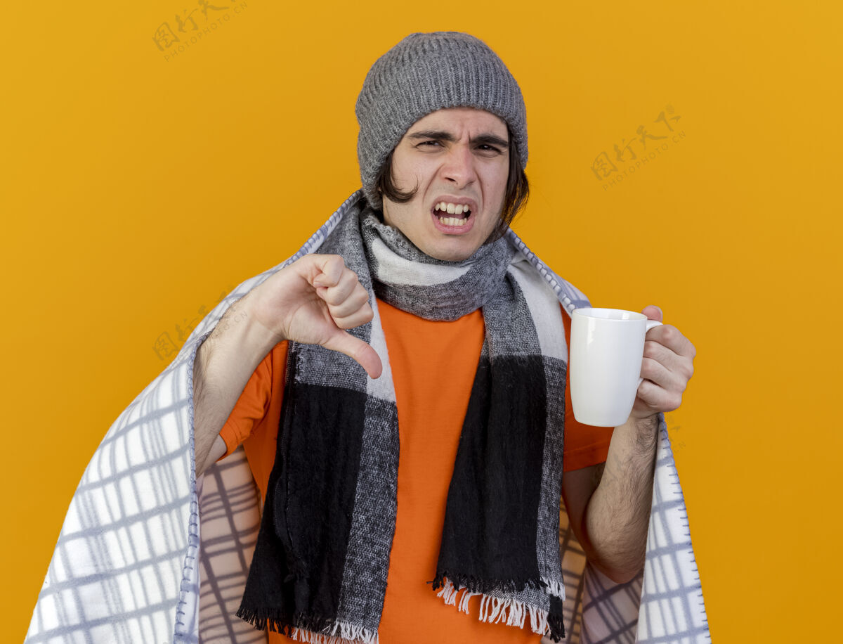 拇指一个戴着冬天帽子 围巾裹着格子布 拿着一杯茶 拇指朝下 被隔离在橙色背景下的未出狱的年轻人疾病拿着格子