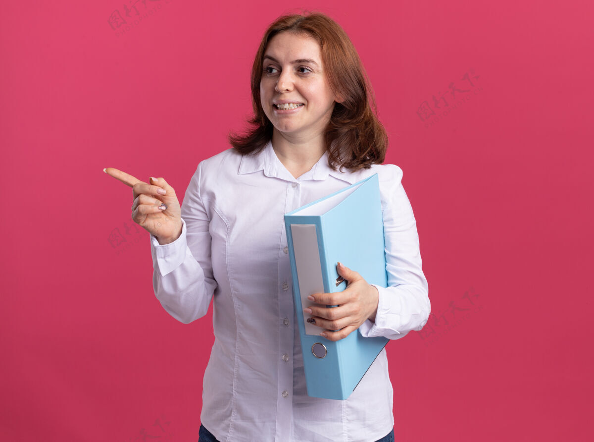 微笑身穿白衬衫的年轻女子拿着文件夹往一旁看 用食指指着一旁 微笑着兴高采烈地站在粉色的墙上衬衫目录女人