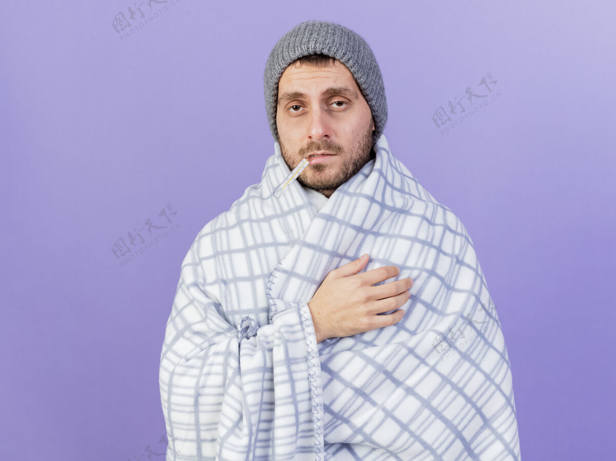 疾病悲伤的年轻病人戴着冬天的帽子 围巾裹着格子布 嘴里叼着温度计 紫色背景上孤立着帽子抱着体温计