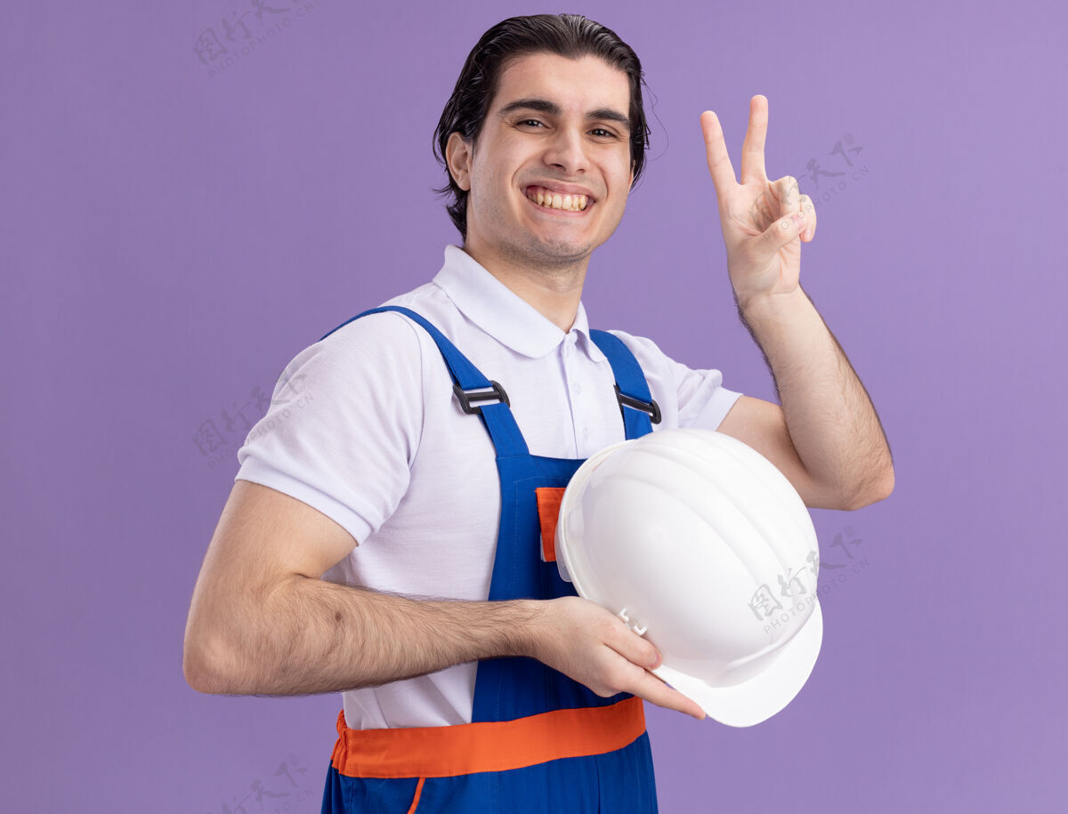 手势身穿建筑制服的年轻建筑工人手持安全帽 自信地微笑着看着前方 紫色的墙壁上立着一个v形标志建筑建筑工人微笑