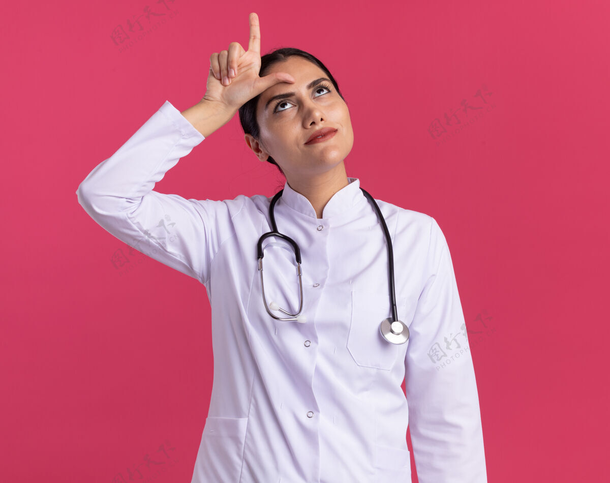 失败者年轻的女医生 穿着医用外套 手持听诊器 脸上带着恼怒的表情抬起头来 站在粉色的墙上做着失败者的手势站立医生手势