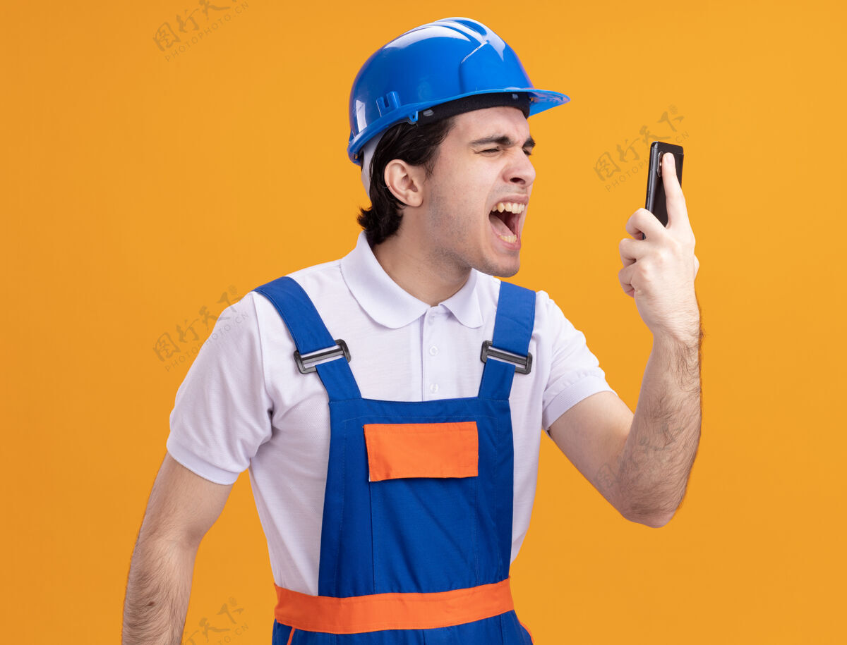 年轻身穿施工制服 头戴安全帽 手持智能手机 表情咄咄逼人的年轻建筑工人站在橙色的墙上大声喊叫制服移动安全