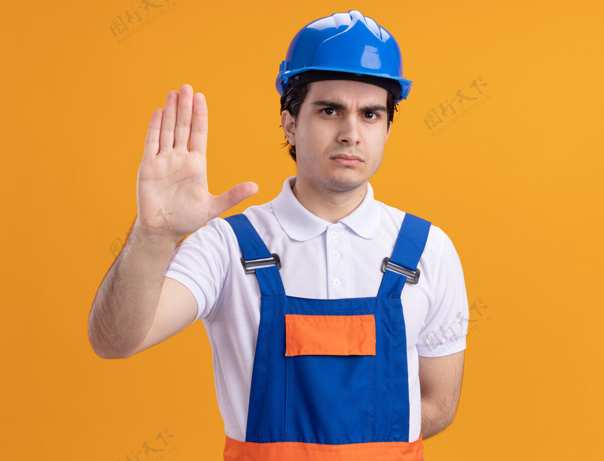 建筑年轻的建筑工人穿着建筑制服 戴着安全帽 严肃地看着前面 用手站在橘色的墙上做着停车的手势手手势停车
