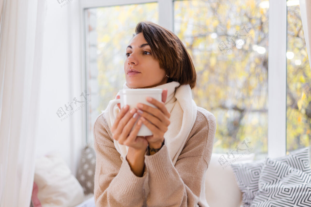放松坐在窗台上喝着热茶喝着咖啡 穿着毛衣和白围巾的女人的温馨肖像室内女人房子