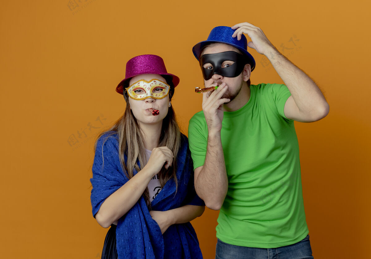帽子年轻快乐的夫妇戴着粉色和蓝色的帽子戴上化装眼罩吹口哨 看起来孤立在橙色的墙上男人派对橙色