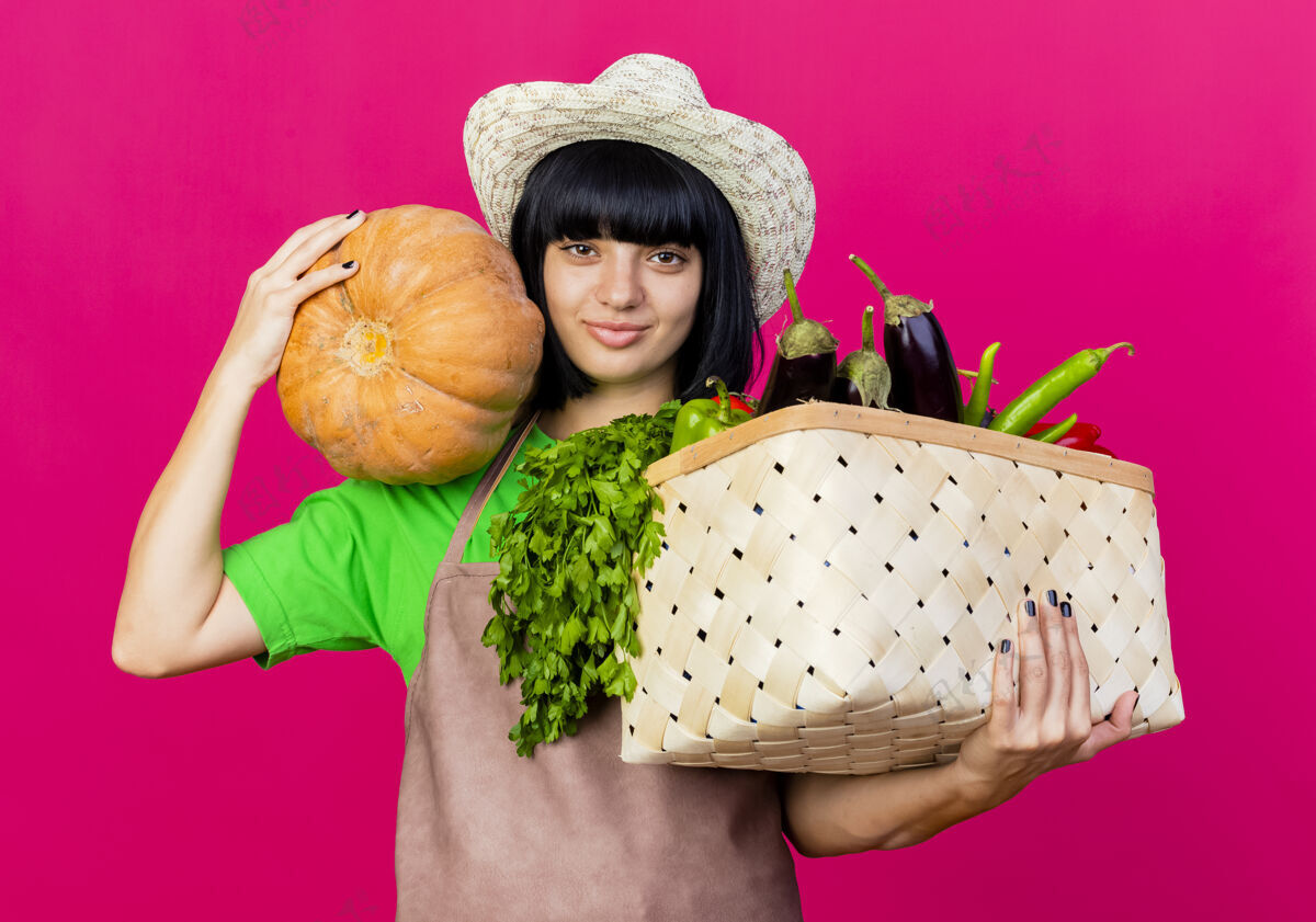 穿高兴的年轻女园丁穿着制服 戴着园艺帽 手里拿着菜篮子和南瓜女粉红色南瓜