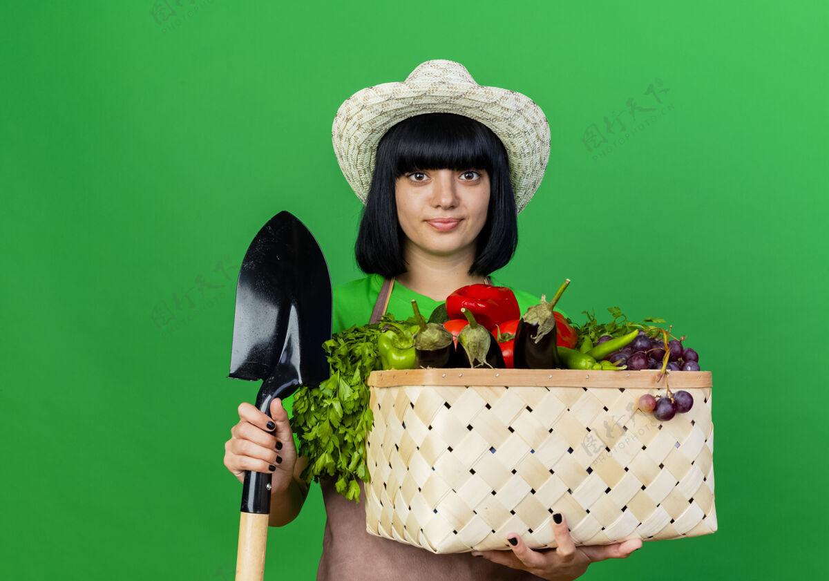年轻高兴的年轻女园丁穿着制服 戴着园艺帽 手里拿着铁锹和菜篮子花园请制服