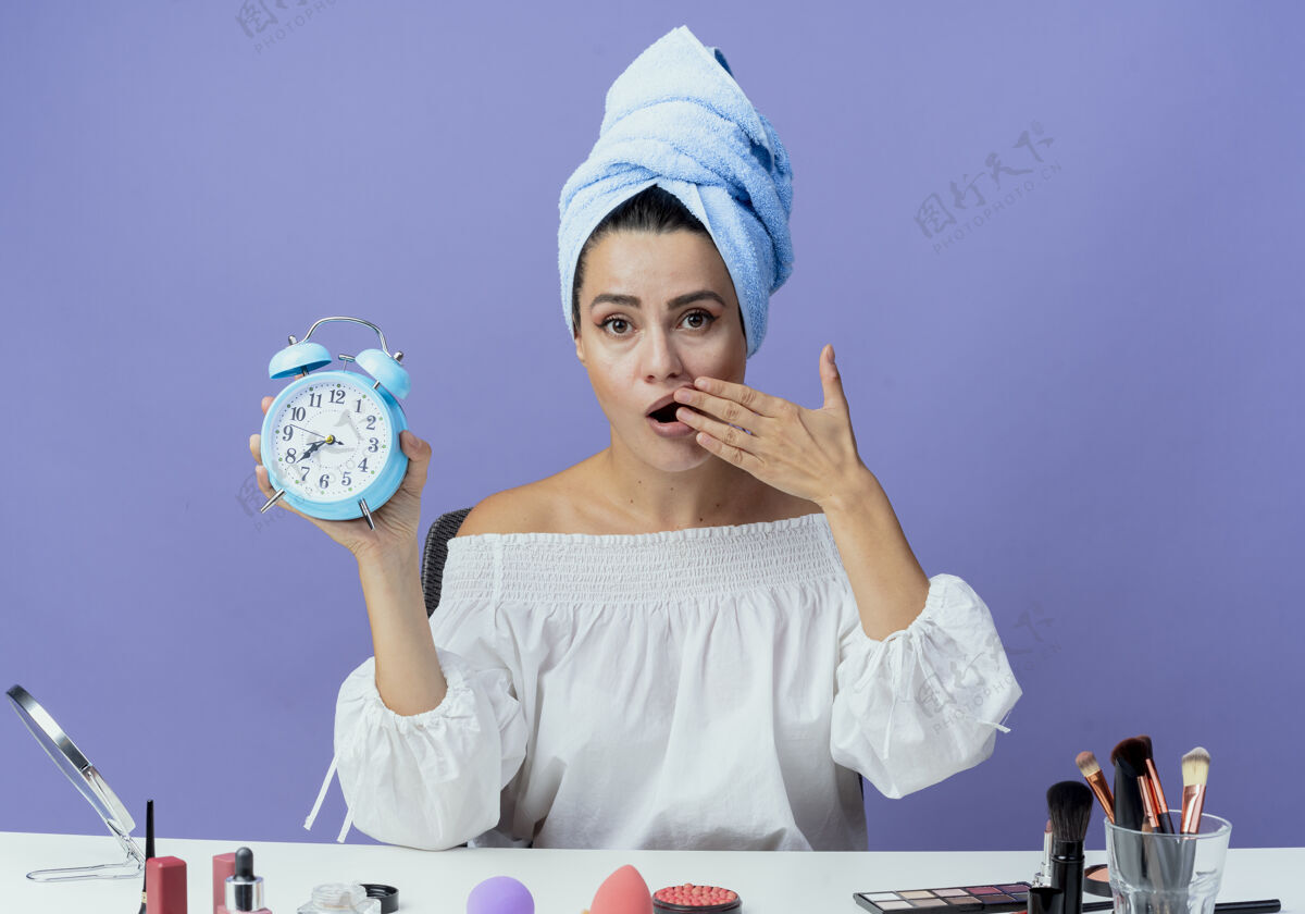 手震惊的美丽女孩裹着毛巾坐在桌子旁 拿着化妆工具 拿着闹钟 手放在嘴上 看起来孤立在紫色的墙上桌子嘴巴美女