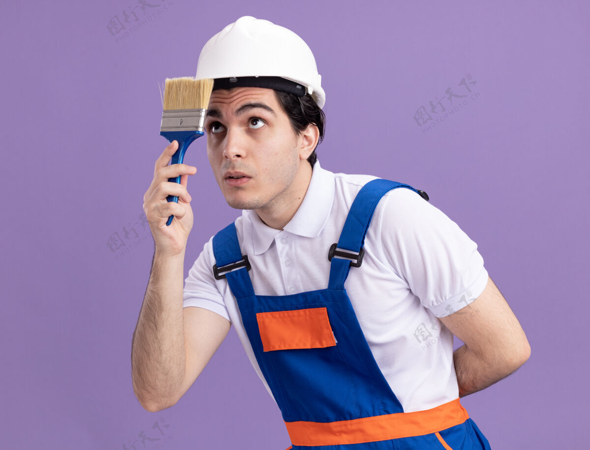 建筑工人年轻的建筑工人穿着建筑制服 戴着安全帽 手里拿着油漆刷 站在紫色的墙壁上好奇地看着它站立制服手持