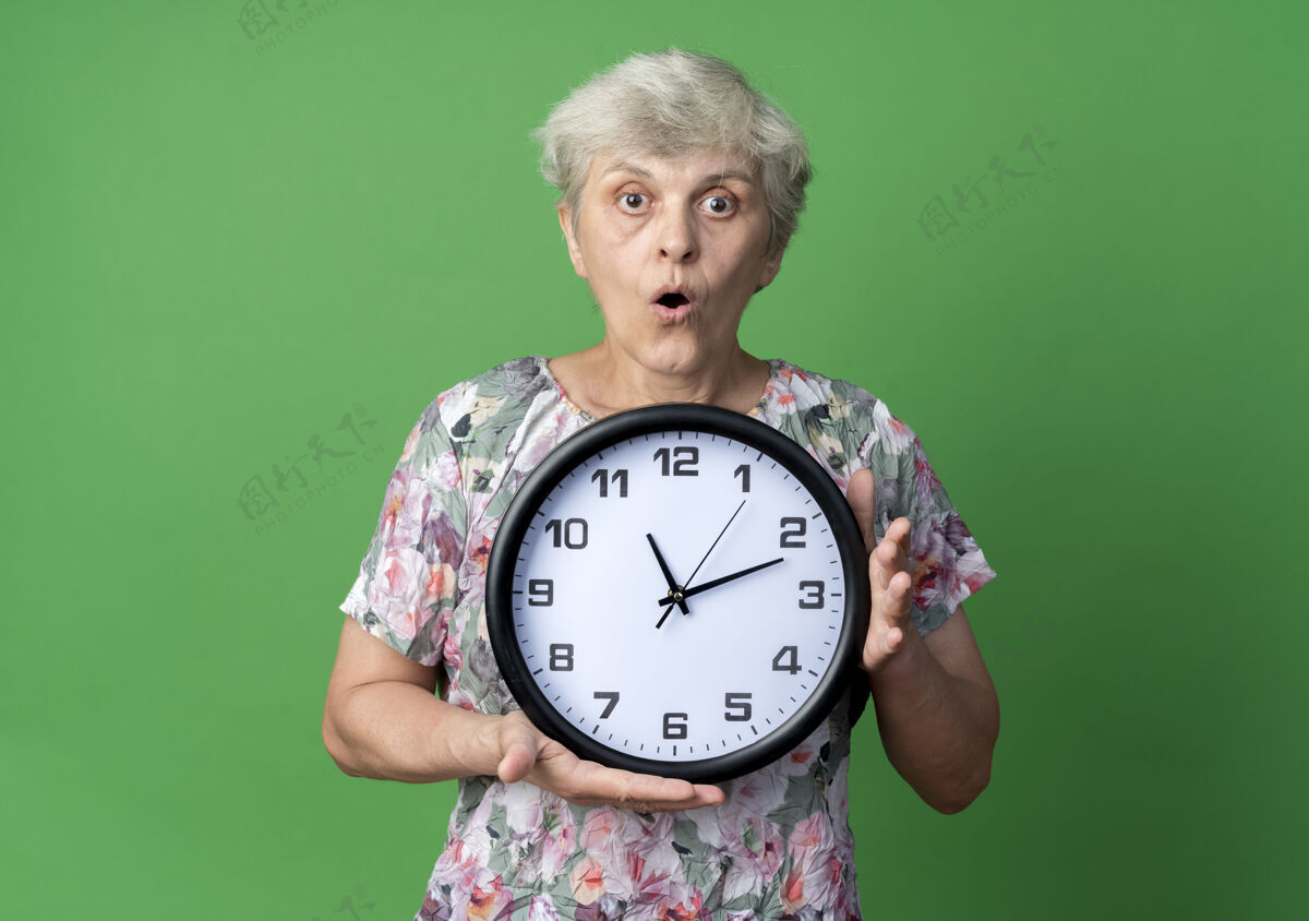 女人惊讶的老妇人把钟孤立地放在绿色的墙上老人老人时钟