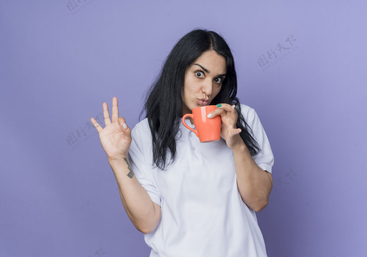 手震惊的年轻黑发高加索人女孩拿着杯子 手势ok手势孤立在紫色的墙上拿着杯子紫色