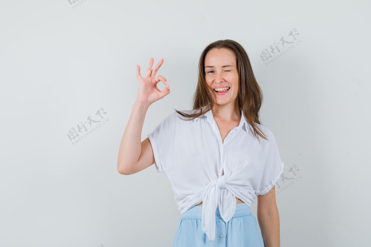 光一个穿着白衬衫和浅蓝色裙子的年轻女人 露出了“好”的手势 伸出舌头 看起来很高兴头发脸肖像