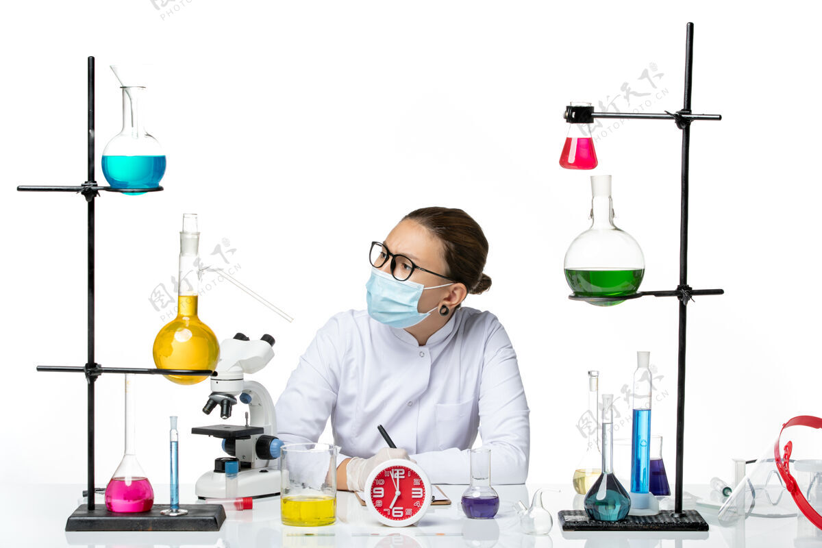 药物前视图：穿着医疗服的女化学家戴着面罩坐着 拿着溶液 在白色背景上写笔记化学家实验室病毒冠状病毒-飞溅套装坐着喷溅