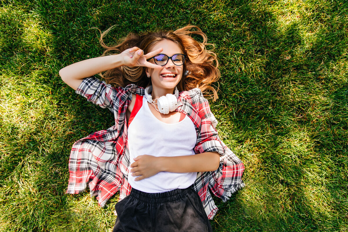 可爱戴着白色耳机的快乐女孩躺在草地上 面带微笑户外俯瞰拍摄的温文尔雅的女人在草坪上发冷年轻放松成人