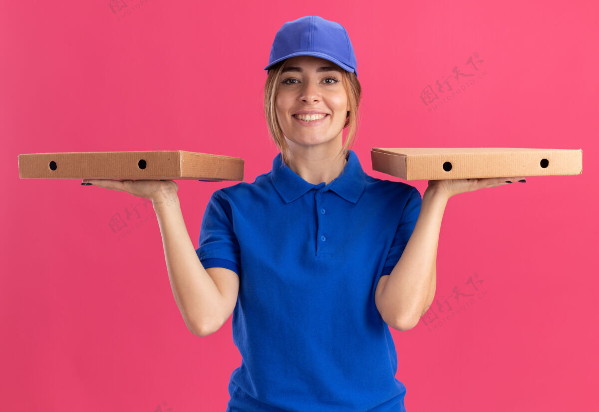 盒子微笑着的年轻漂亮的送货女孩 穿着制服 双手捧着披萨盒 粉色的漂亮披萨姿势