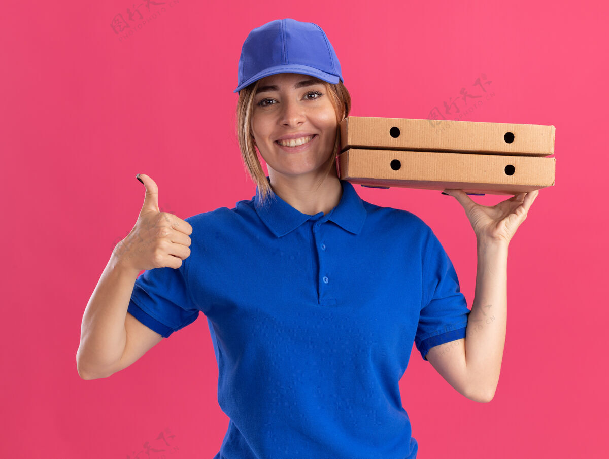 送货微笑着的穿着制服的年轻漂亮的送货女孩竖起大拇指 把比萨饼盒贴在粉色的脸上盒子漂亮制服