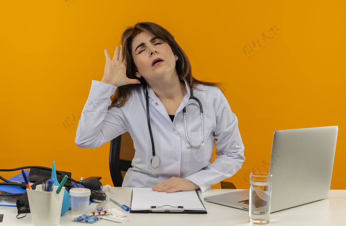 医疗疼痛的中年女医生穿着医用长袍和听诊器坐在办公桌旁 用医疗工具 剪贴板和笔记本电脑摸着头痛的头疼痛橙色桌子