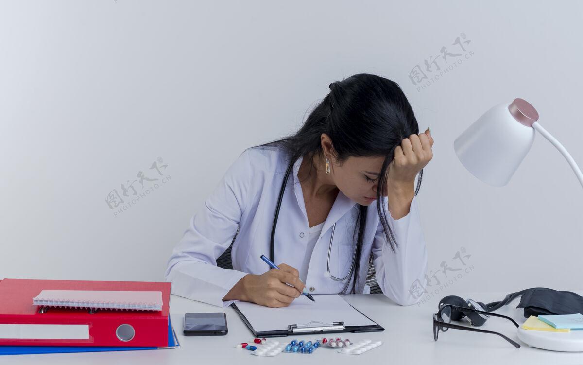 笔疲惫的年轻女医生穿着医用长袍 手持听诊器坐在办公桌旁 手持医疗工具 手放在头上 闭着眼睛 与世隔绝累了医生女人