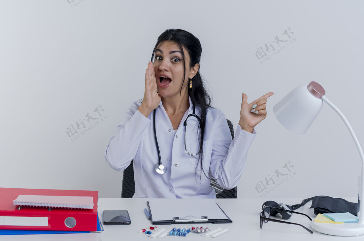 穿着令人印象深刻的年轻女医生穿着医用长袍和听诊器坐在办公桌旁 用医疗工具指着旁边 低声说话人指点年轻人