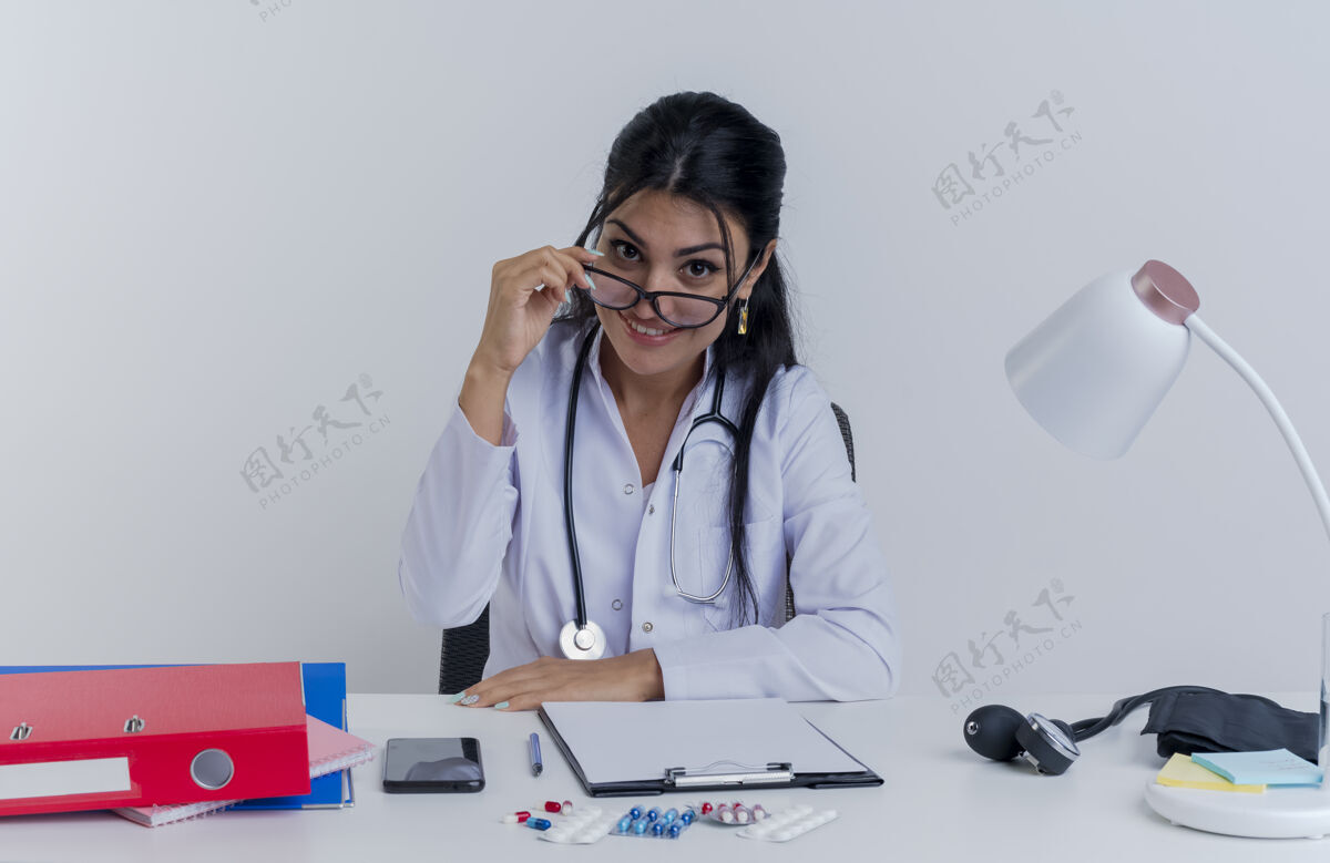 工具面带微笑的年轻女医生 穿着医用长袍 戴着听诊器和眼镜 坐在桌子旁 拿着医疗工具看着 把手放在桌子上 抓起眼镜穿人年轻人
