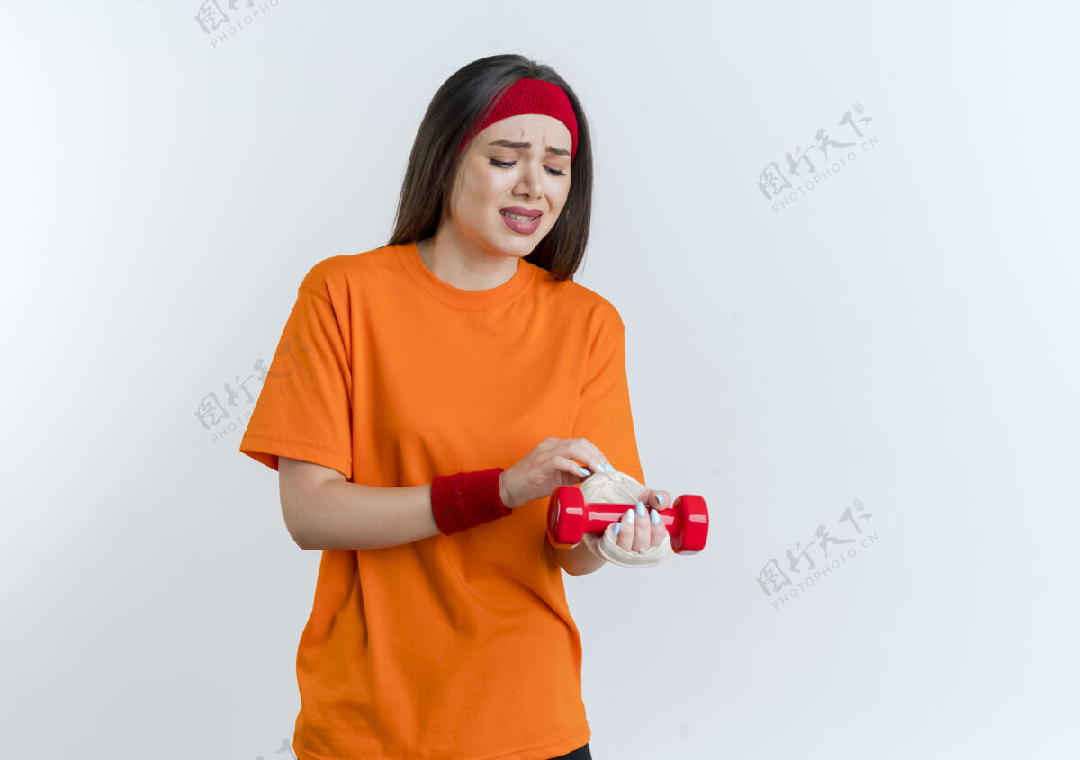疼痛疼痛的年轻运动女性戴着头带和手环举着哑铃抚摸着 看着裹着绷带的受伤手腕抱着手腕受伤
