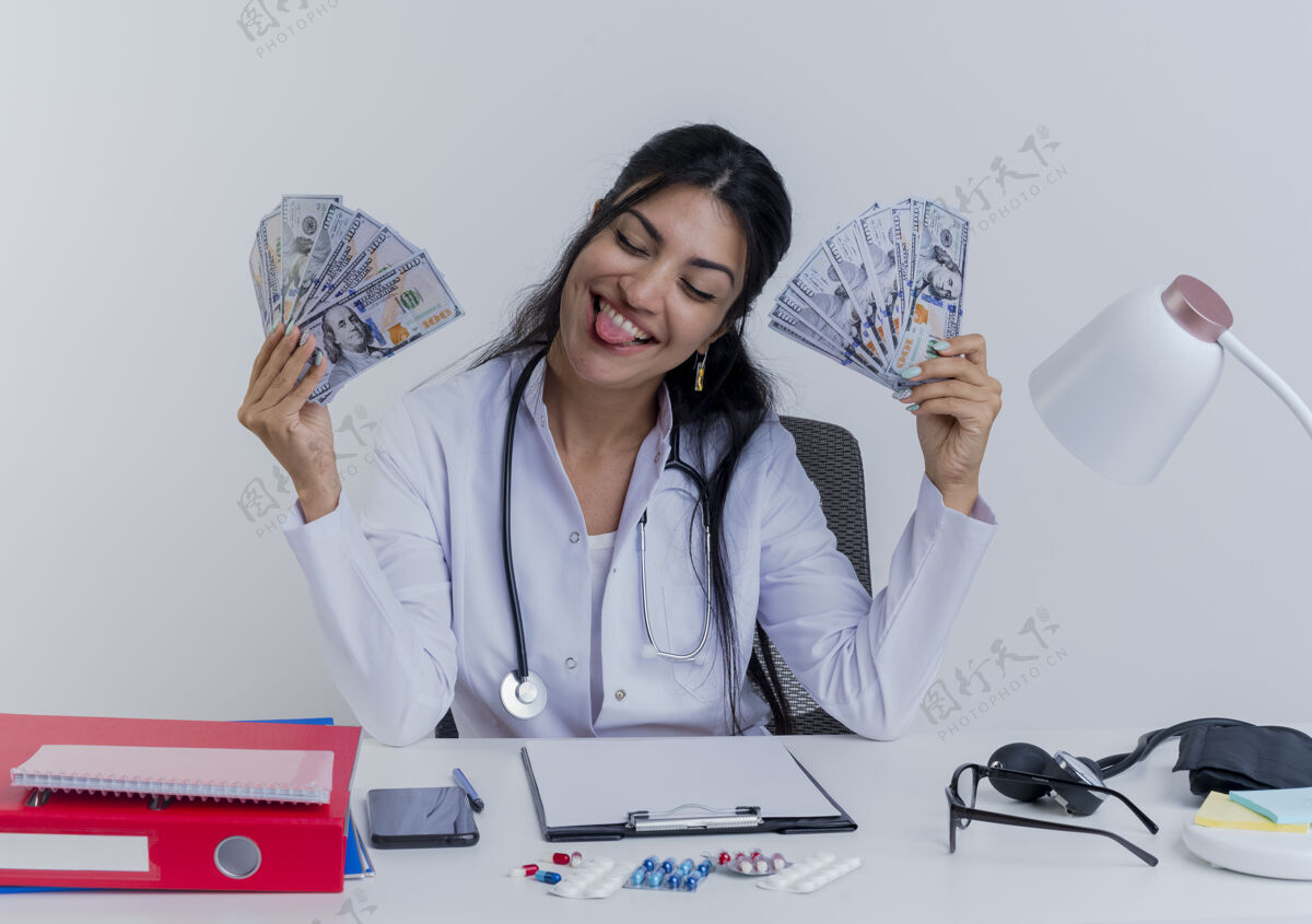 成人顽皮的年轻女医生穿着医用长袍和听诊器坐在办公桌旁 手里拿着医疗工具 手里拿着钱 闭着眼睛 露出舌头抱着穿女人