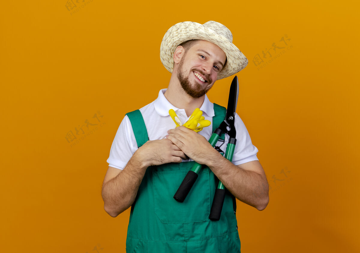 年轻快乐的年轻英俊的斯拉夫园丁穿着制服戴着帽子拿着园艺手套和修剪看起来孤立帅哥男人园艺