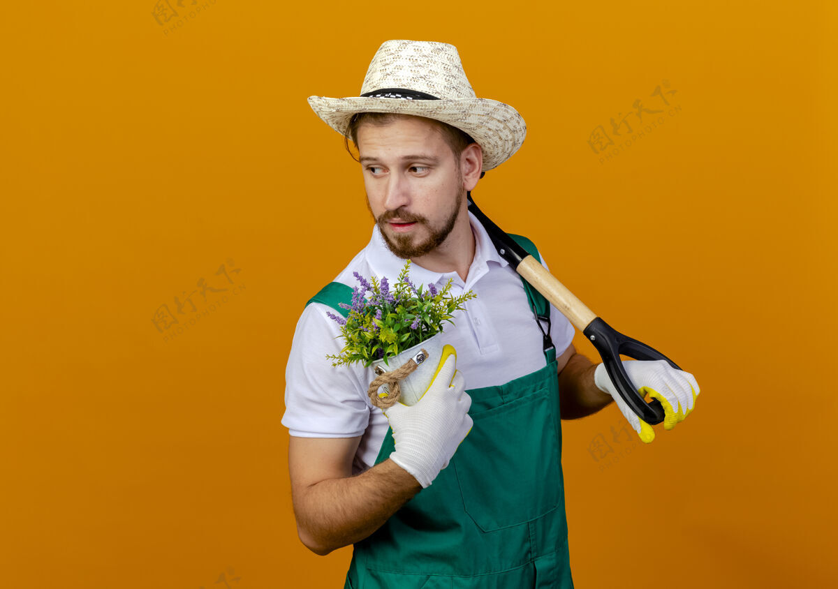 年轻自信的年轻英俊的斯拉夫园丁 穿着制服 戴着帽子和园艺手套 肩上扛着铁锹 手里拿着花盆 看着孤立的一面黑桃持有手套