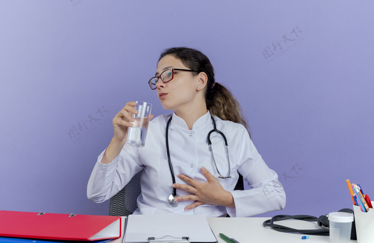胸部年轻的女医生穿着医用长袍和听诊器坐在办公桌旁 手持医疗工具把头转向一边 手放在胸前 手里拿着一杯水女人成人长袍