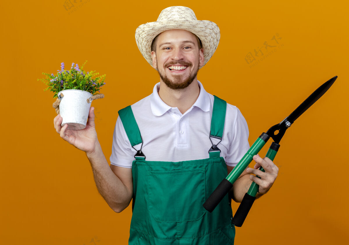 男人快乐的年轻英俊的斯拉夫园丁 穿着制服 戴着帽子 手里拿着花草和修剪机 看上去与世隔绝修剪者橙色年轻