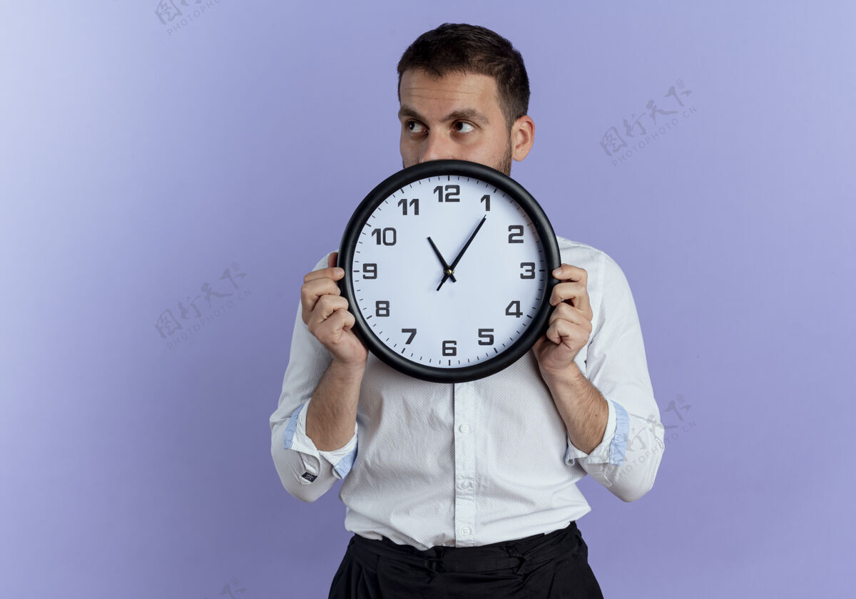坚持自信的帅哥拿着时钟看着紫色墙上孤立的一面紫色男人时钟