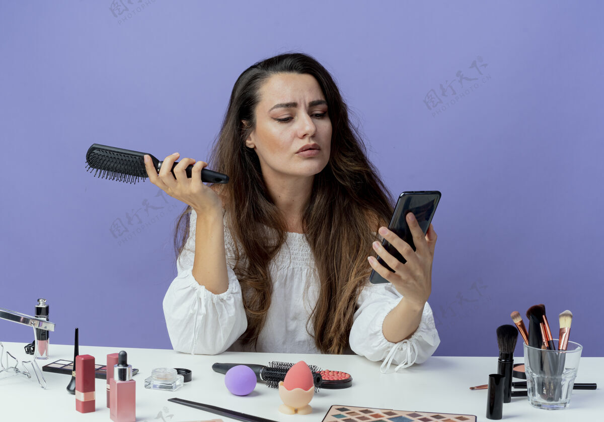 抱着自信美丽的女孩坐在桌子旁 拿着化妆工具 拿着梳子 看着紫色墙上的电话电话自信工具